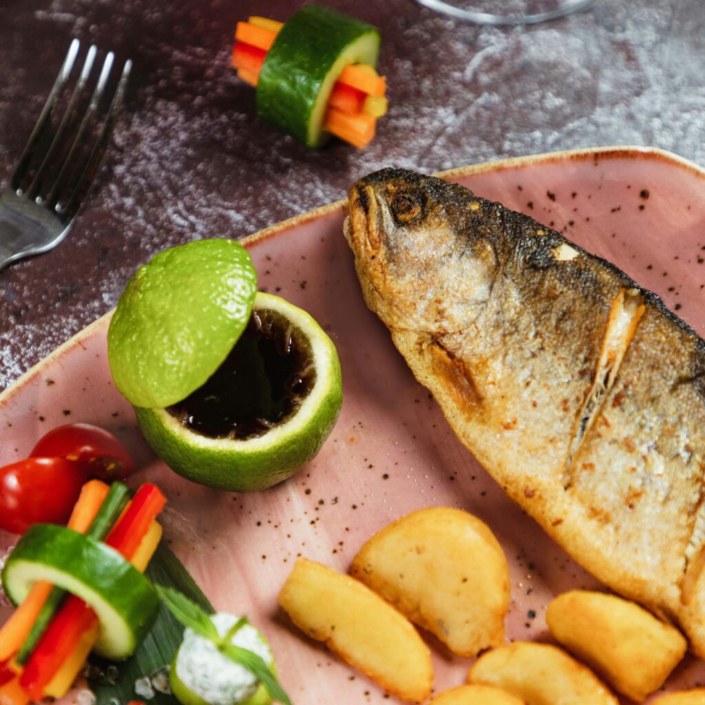 Delicious grilled whole fish served with fresh vegetables and lime on a pink plate.